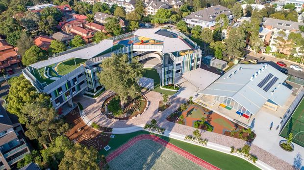Aerial view of a large contemporary school or institutional campus in a leafy suburban setting. The complex consists of several interconnected low-rise buildings with flat roofs, extensive glazing, rooftop play or recreation areas, paved courtyards, landscaped spaces, and prominent outdoor sports courts, including a tennis or multi-purpose court in the foreground.