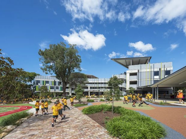 Exterior view of a contemporary multi-storey educational or institutional campus with students walking through landscaped courtyards and along a brick-paved path. The buildings feature extensive glazing, white and grey cladding, vertical coloured facade accents, broad projecting roof canopies, and a prominent elevated flat canopy. A running track and covered entry areas are visible, with bright blue sky and mature trees surrounding the complex.
