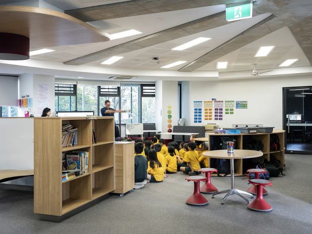An open-plan school learning commons or library with children seated on the carpet in a group activity. The space features low timber bookcases and shelving, round tables with colorful pedestal stools, educational wall displays, large windows with horizontal blinds, carpet flooring, and a contemporary exposed-services ceiling with recessed lighting. A staff member and reading materials are visible in the background.
