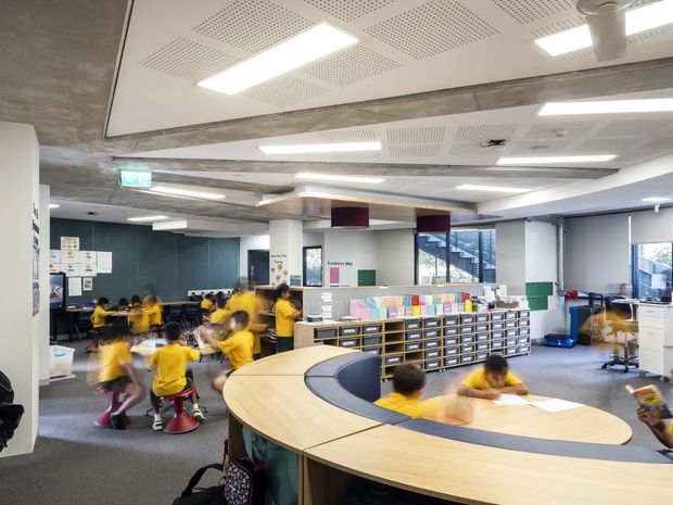 A bright, open-plan school learning and activity space with children seated and moving around curved built-in timber workstations. The room includes extensive low storage and pigeonhole cabinetry, collaborative tables, display materials, carpeted flooring, acoustic perforated ceiling panels, exposed concrete structural beams, and large windows. The atmosphere is energetic and functional, with a colourful educational fit-out.