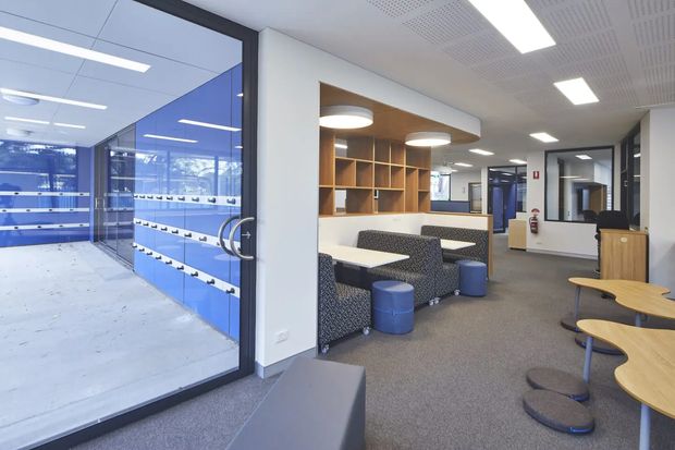 A bright contemporary commercial interior, likely a school, office, or institutional facility, showing a carpeted breakout lounge beside a glazed corridor or wet-area space. The lounge contains patterned upholstered booth seating, white tables, blue cylindrical stools, timber shelving, and curved timber tables. The space has white walls, a suspended acoustic perforated ceiling with recessed linear lighting, dark-framed internal glazing, and a fire extinguisher visible in the corridor.