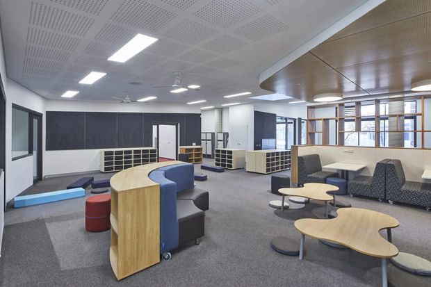 A spacious contemporary commercial interior, likely a collaborative workplace or education lounge, with open-plan seating areas, curved timber-panelled furniture, upholstered lounge chairs and sofas, small round tables, built-in shelving, grey carpet, dark feature walls, and a suspended acoustic ceiling with integrated LED lighting. Glazed partitions and timber-framed screening define areas toward the rear.