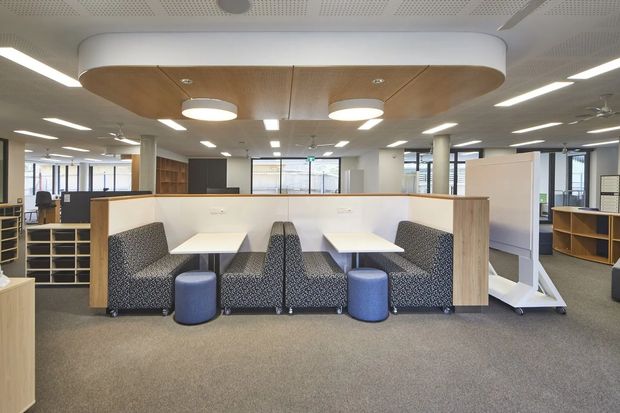 A bright contemporary open-plan commercial workspace with paired booth-style workstations, white desks, patterned upholstered seating, and blue ottomans. Timber-toned partitions and shelving define the workspace, while recessed linear lighting, ceiling fans, acoustic perforated ceiling panels, and a large curved timber ceiling feature create a polished office fit-out.