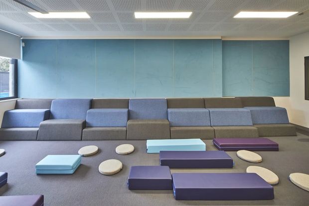 A contemporary commercial lounge or informal waiting area with tiered modular upholstered seating against a blue acoustic-panel wall. Low geometric ottomans and tables in blue, purple, beige, and grey are arranged across a broad carpeted floor. The space has a perforated suspended ceiling with rectangular fluorescent-style light panels and strong daylight from side glazing.