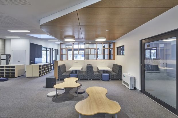 Contemporary commercial interior showing an open lounge and collaborative breakout area. The space includes upholstered booth seating, small organic-shaped timber coffee tables, grey carpet, white cabinetry, glazed partitions, and a prominent curved timber ceiling feature. Recessed and pendant lighting create a bright, professional workplace atmosphere.