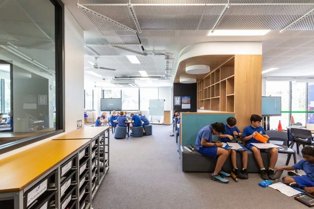 A bright contemporary school learning commons or collaborative study area, viewed from an open circulation zone. The space includes long timber-topped work counters with drawer storage, glass-partitioned rooms, group tables, lounge seating, integrated shelving, and students working in small groups. A perforated suspended ceiling, exposed services, large windows, and colourful furnishings create an open, functional educational interior.