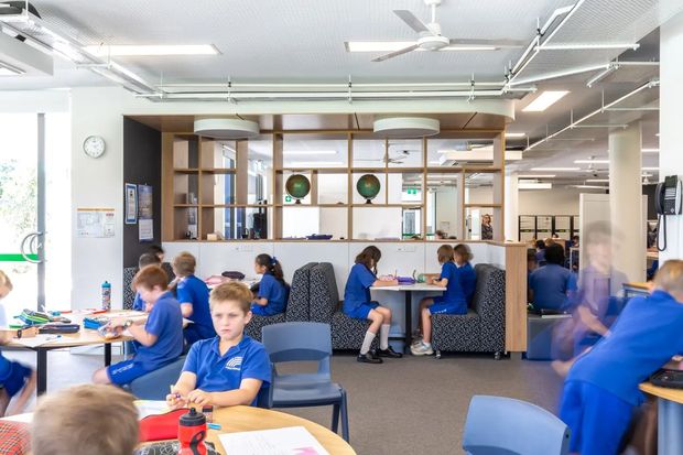 A bright, open-plan school or institutional learning commons with students seated at collaborative tables and booth seating. The space features a contemporary timber-framed open shelving and room-divider structure, extensive glazing, carpeted flooring, white acoustic ceiling panels, exposed services, ceiling fans, and abundant daylight. Blue school uniforms and furnishings provide strong colour accents. The image primarily depicts a commercial or educational interior rather than a residential room.