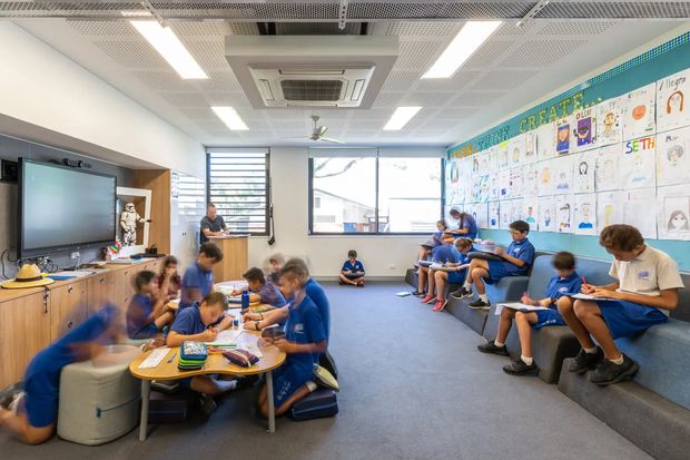 A bright, contemporary school classroom or learning commons occupied by students working in small groups and seated along tiered upholstered bench seating. The room features a large wall-mounted display, extensive student artwork, blue carpet, timber-toned built-in joinery, generous windows, and a perforated acoustic ceiling with linear lighting and a central air-conditioning unit.