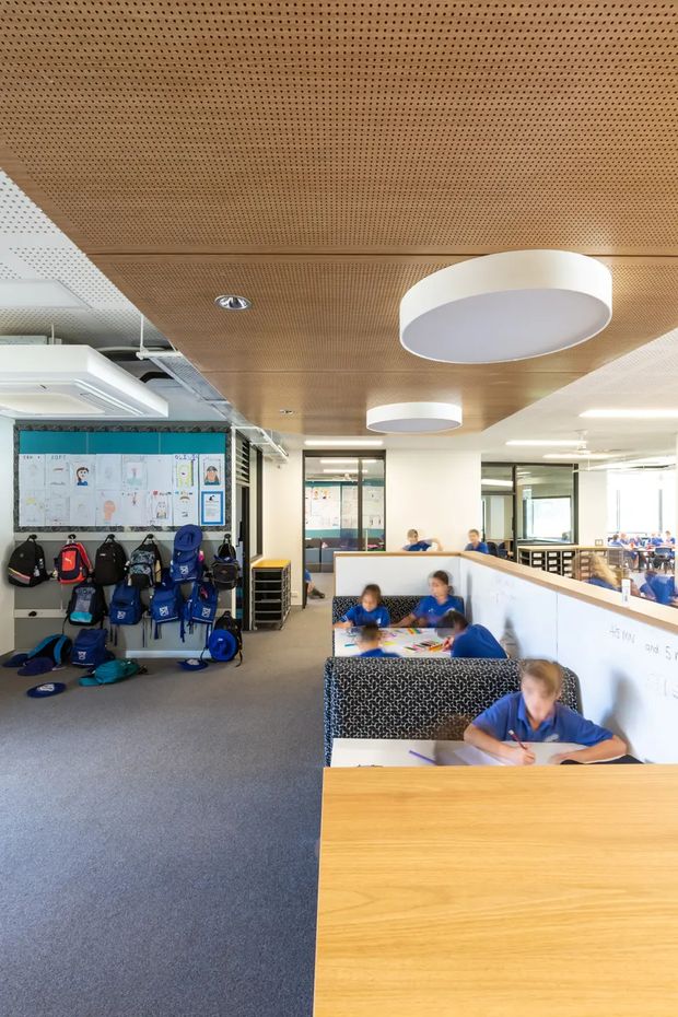 Bright open-plan school learning area with students working at built-in study booths and desks. The space features a warm perforated timber acoustic ceiling, carpeted flooring, white partitions and cabinetry, large windows, circular pendant lighting, and a designated wall-mounted storage area for school bags and equipment.
