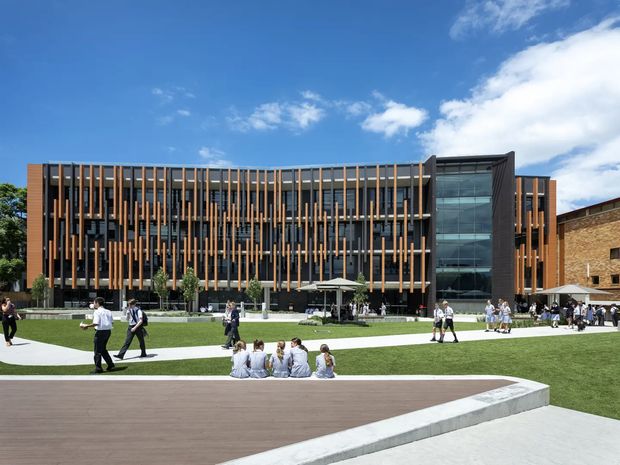 Wide exterior photograph of a contemporary multi-storey institutional building facing a landscaped campus courtyard. The facade combines extensive dark glazing with vertical timber-toned fins and dark structural framing, plus a prominent glazed circulation or entry tower. Students and pedestrians occupy the lawn and paved walkways in front, with a timber-toned deck and concrete edge in the foreground under a bright blue sky.