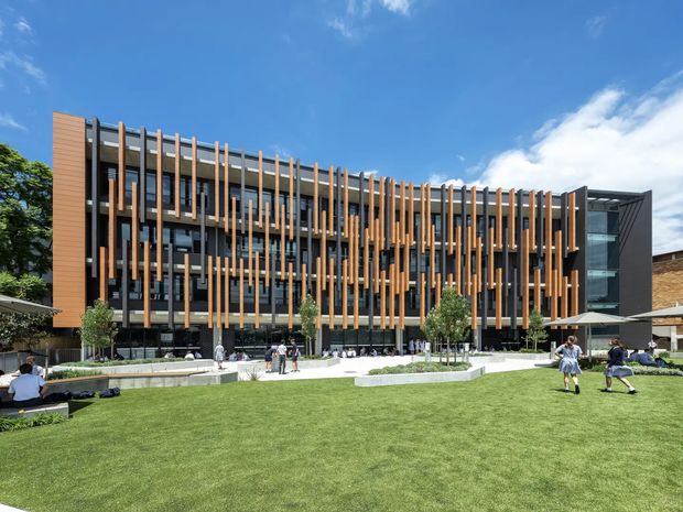 A contemporary multi-storey institutional or commercial building viewed from a landscaped lawn. The facade combines extensive dark-framed glazing with a strong rhythm of vertical timber-toned fins and darker structural elements. The building opens onto a bright courtyard-like lawn with paved seating areas, umbrellas, small trees, and people using the space. A projecting glazed corner and angular dark side volume add visual emphasis.