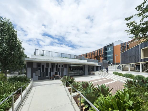 Exterior view of a contemporary multi-level institutional or commercial complex with landscaped courtyards, accessible ramps, broad paved terraces, dark tiled walls, orange vertical facade elements, extensive glazing, and rooftop railings. The image focuses on the building frontage and outdoor circulation spaces.