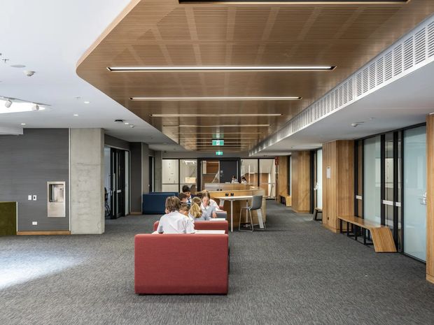 A contemporary open-plan commercial interior with collaborative lounge seating, a central kitchenette or café-style breakout area, and glazed meeting rooms along the sides. The space features a warm timber slatted ceiling with integrated linear lighting, dark carpet flooring, exposed concrete and grey wall finishes, timber joinery, and extensive internal glazing. Several people are seated in the informal workspace.