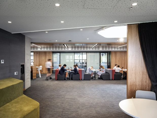 A contemporary open-plan commercial collaboration and breakout area with groups of people seated in lounge chairs around small tables. The space features warm timber wall panelling and columns, grey carpet, dark charcoal wall and curtain surfaces, a patterned acoustic ceiling with recessed downlights, extensive internal glazing, and a large curved skylight-like ceiling feature. Tiered green upholstered seating is visible in the foreground, with a white round table and chairs at right.