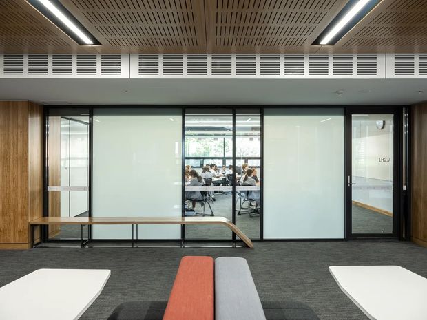 Contemporary commercial interior showing a corridor or breakout area with built-in timber wall panelling, dark carpet flooring, black-framed glazed office partitions, and a slatted timber acoustic ceiling with linear lighting. A meeting or collaborative workspace is visible through the central glazing, with colourful upholstered benches in the foreground.