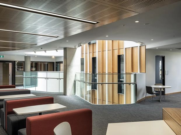 A contemporary commercial interior, likely a reception or lounge area, with grouped upholstered seating, small tables, glass balustrades, and a prominent curved timber-clad central feature enclosing a stair or circulation zone. The space has a patterned acoustic ceiling, recessed and linear lighting, carpet flooring, exposed concrete columns, and a neutral grey, white, timber, and muted red palette.