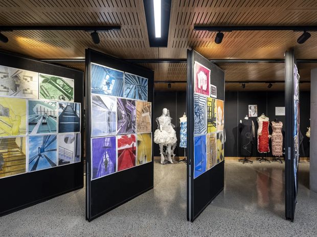 A contemporary exhibition or gallery interior displaying fashion garments and illustrated design boards on freestanding black partition panels. The space has a warm slatted timber ceiling, integrated linear lighting and spotlights, dark walls, polished speckled flooring, and a mannequin display in the background.