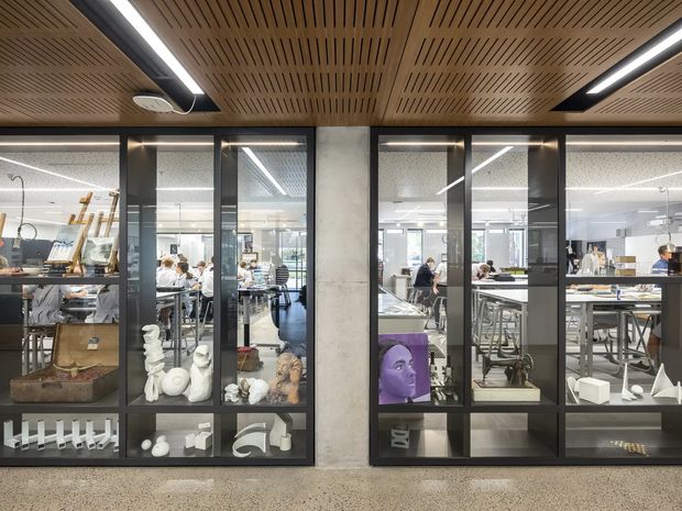 A contemporary commercial interior viewed through black-framed glazed partitions. The foreground features built-in display cabinetry containing sculptural objects and models, while the glass reveals a bright open-plan studio or workshop with desks, stools, shelving and people working. A timber slatted acoustic ceiling with integrated linear lighting spans the space, and the floor appears to be polished concrete or terrazzo.