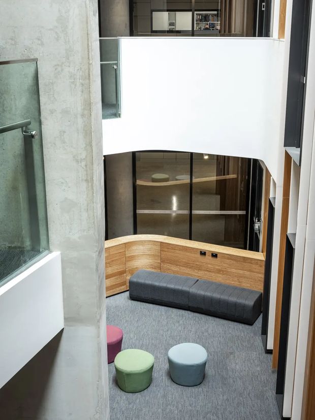A contemporary multi-level interior atrium or lounge viewed from above, featuring a curved timber-clad built-in seating wall, dark upholstered bench, small rounded stools, grey carpet, exposed concrete, glass balustrades, and a large interior glazed partition. A white curved balcony edge and vertical timber detailing frame the double-height space.