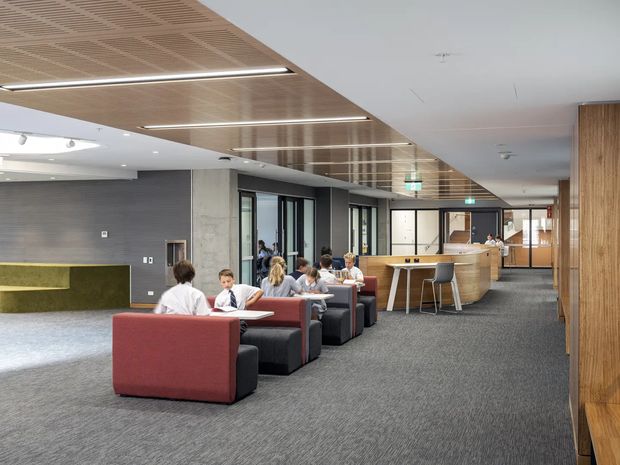 A modern commercial interior resembling a library, university common area, or corporate lounge. The open-plan space features clustered red and dark upholstered lounge seating, a long curved timber reception or service counter with stools, grey carpet, concrete and dark grey wall finishes, extensive glazing, and a suspended timber slatted ceiling with linear integrated lighting. People are seated and working informally throughout the space.