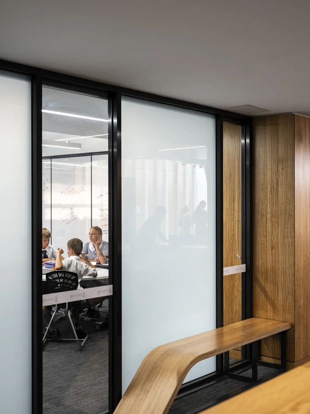 Contemporary commercial office interior with black-framed glazed partitions, translucent privacy film, a meeting room visible through the glass, and warm timber wall panelling with an integrated curved timber bench. The space has dark carpet flooring, recessed ceiling services, and a refined corporate fit-out.