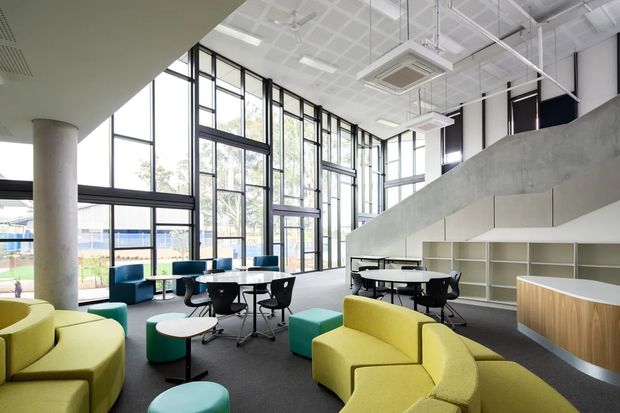 A bright, expansive contemporary commercial interior with a double-height volume, full-height black-framed glazing, exposed concrete stair or mezzanine structure, and an open collaborative lounge or workspace. The room contains curved lime-yellow upholstered seating, teal ottomans and chairs, small round tables, black task chairs, communal tables, and built-in pale timber and white shelving. A patterned suspended ceiling with exposed services and large linear windows gives the space an airy institutional or office character.