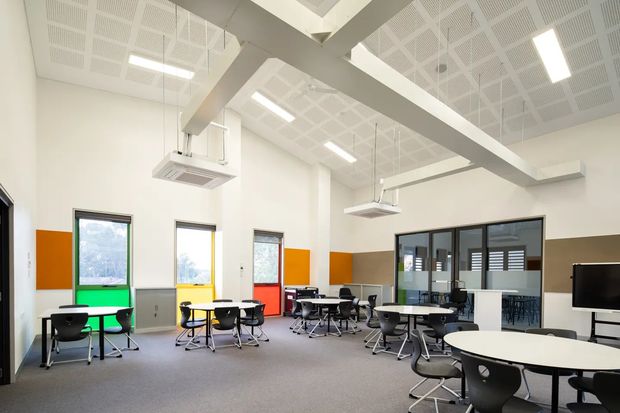 A bright, contemporary commercial classroom or collaborative learning space with multiple round tables and black chairs arranged throughout. The room has a high exposed structural ceiling with suspended acoustic panels, linear LED lighting, suspended air-conditioning units, large windows with vivid orange, green, yellow and red lower wall panels, and a glazed partition enclosing an adjoining workspace or meeting room. Neutral carpet and white walls create a clean, functional environment.