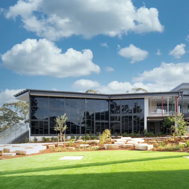 A modern low-rise building viewed from landscaped grounds, featuring extensive dark reflective floor-to-ceiling glazing, a flat roof with broad eaves, light rendered or concrete walls, and a covered upper-level balcony with slender red vertical accents. The foreground includes a bright lawn, paved paths, planted beds, and large pale stone seating blocks. The image is an exterior architectural photograph under a blue sky.
