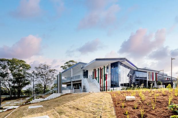 A contemporary multi-storey institutional or commercial building set on a landscaped sloping site. The building features angular low-pitched roof forms, extensive glazing, white and grey facade panels, vertical white fins, and colourful red and green vertical accents. External stairs, accessible ramps, planted garden beds, and open circulation areas are visible.