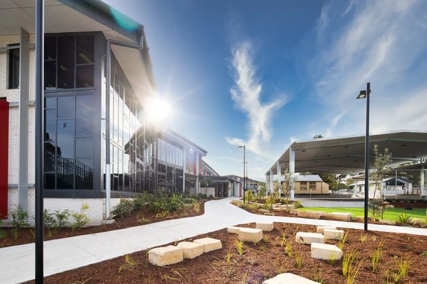 Exterior view of a contemporary institutional or commercial complex with glazed multi-storey facades, covered outdoor structures, paved pedestrian paths, landscaped garden beds, and integrated outdoor seating. The scene is bright and open, with clean-lined architecture and a flat-roofed canopy prominent on the right.