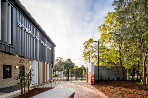 Exterior view of the Schofields Public School building, featuring dark vertical metal cladding, pale brickwork, a projecting roof edge, signage, security fencing and a paved pedestrian entry courtyard surrounded by established trees and landscaped garden beds.