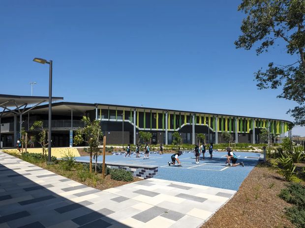 Exterior view of a contemporary public or commercial recreation building with a long curved facade, deep roof canopy, and vertical lime-green and dark-grey facade elements. A blue outdoor basketball court and paved pedestrian forecourt occupy the foreground, with landscaped planting around the site.