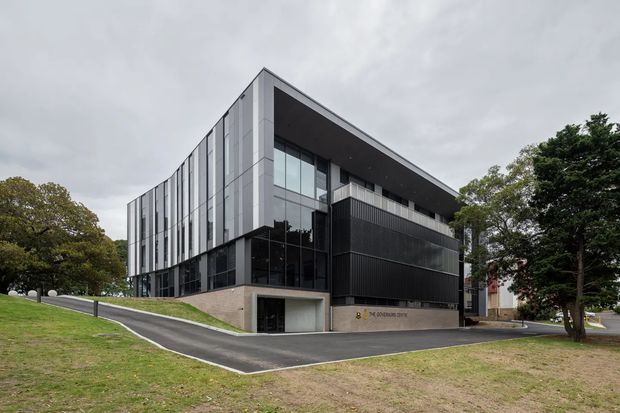 A modern multi-storey commercial building set within a landscaped, gently sloping site. The building has a sharply angled roofline, extensive dark-framed glazing, vertical light-grey facade panels, dark ribbed cladding, and a recessed ground-level entry or parking area. Overcast conditions and mature trees surround the property.