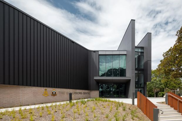 Exterior architectural photograph of The Governors Centre, a contemporary institutional building. The composition features a long dark vertically ribbed facade, pale masonry base with signage, a glazed entrance volume, and sharply projecting grey metal-clad forms. A landscaped gravel forecourt and stair access occupy the foreground beneath a partly cloudy sky.