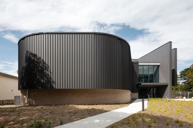 Contemporary multi-part building exterior featuring a prominent rounded volume clad in dark vertical ribbed panels, set above a curved textured masonry base. A contrasting angular wing has large glazed openings and smooth grey panel cladding. A pale concrete pathway leads through a sparsely landscaped foreground under a partly cloudy sky.