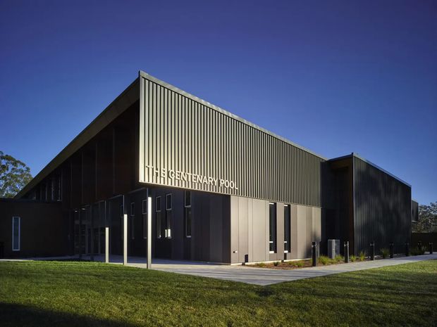 A contemporary single-storey institutional building photographed from the exterior. The structure has a strongly geometric form with a tall pitched or sloping roof volume, dark vertical metal cladding, extensive ribbed screening, narrow vertical windows, and a recessed glazed entrance. A sign reading “Centenary Pool” is mounted on the facade. The building sits beside a paved entry path and landscaped lawn under a clear blue sky.
