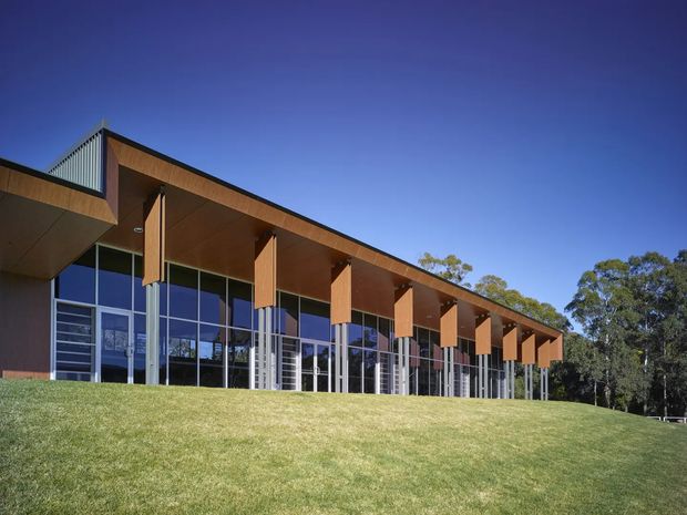 Exterior view of a low, modern pavilion-like building set on a gently sloping lawn. The facade features extensive floor-to-ceiling glazing, slender metal framing, a deep projecting roof canopy, and repeated timber-clad vertical fins supporting the eaves. A small ribbed metal volume rises above part of the roofline, with mature trees visible behind the building.