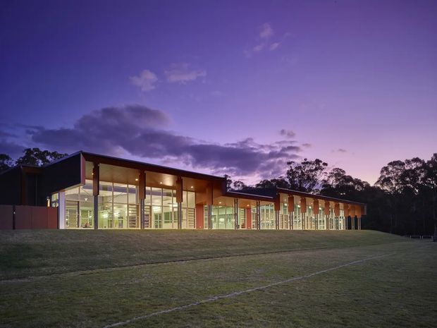 A low, elongated modern building photographed at dusk, set beside a broad grassed field. The facade features extensive floor-to-ceiling glazing, warm interior lighting, slender vertical structural elements, and dark projecting roof planes, with mature trees forming the background.