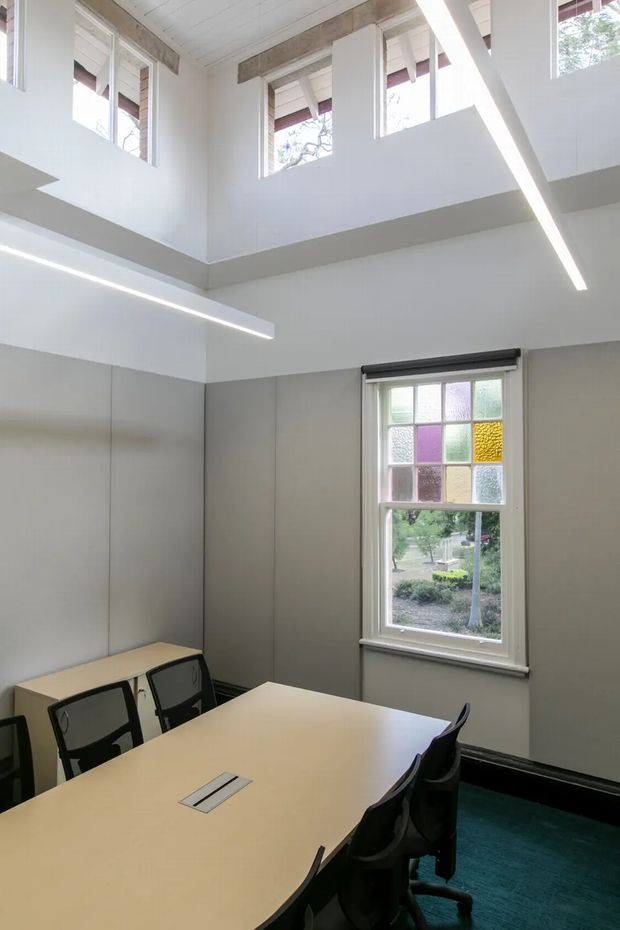 A bright double-height meeting or collaboration room with pale grey wall panels, white upper walls and ceiling, exposed timber beams, high clerestory windows, and contemporary linear pendant lighting. A large light-coloured conference table is surrounded by black office chairs. The lower window includes a colourful stained-glass-style grid, while blue carpet covers the floor.