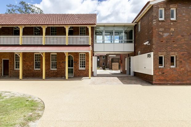 Exterior view of a heritage-style brick complex with a two-storey verandah-fronted building on the left and a taller brick building on the right, linked by a contemporary glazed passage. The buildings feature red-brown brickwork, tiled pitched roofing, timber verandah posts and balustrades, multi-pane windows, and a paved courtyard or access area in the foreground.