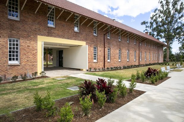 Exterior view of a long, low-rise brick building with a pitched terracotta-tiled roof, exposed timber eaves and repetitive narrow windows. A large recessed entrance punctuates the facade, opening onto landscaped gardens and concrete pedestrian paths.