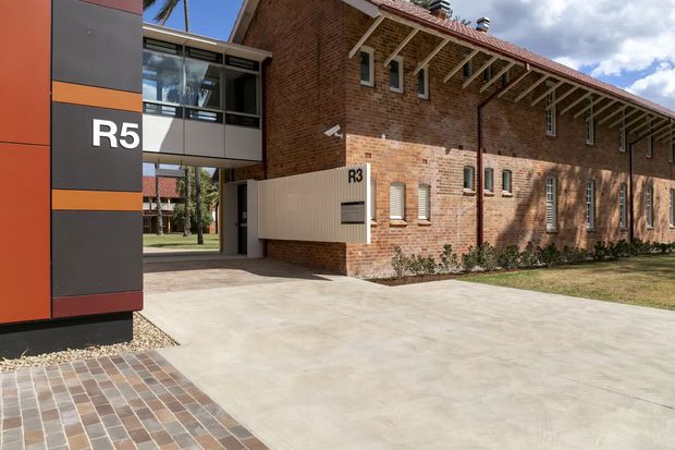 Exterior architectural photograph of a low-rise institutional or commercial complex. A contemporary dark grey and orange-clad entry volume marked “R5” sits beside a renovated red-brick building marked “R3”. The brick structure has a long tiled pitched roof, exposed projecting rafters, narrow vertical windows, and a covered passage connecting the buildings. Concrete paving and a patterned brick walkway occupy the foreground, with landscaped grounds visible beyond.