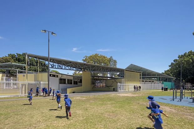 Exterior view of a modern primary school campus with low-rise pale yellow buildings, extensive steel-framed covered walkways and pitched canopy roofs. Children are playing across a grassy sports area in front, with fenced courts and outdoor exercise equipment visible to the right. The building signage identifies it as a public school. Bright, clear daytime setting.