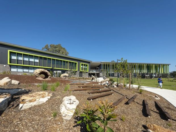 A low-rise contemporary institutional or commercial building viewed from a landscaped outdoor area. The building has dark grey horizontal cladding, extensive floor-to-ceiling glazing, vivid lime-green window and facade frames, and a low-pitched roof. A covered link connects the main wings, with vertical screening elements along the right-hand facade. The foreground contains native-style landscaping, large exposed rocks, timber edging/logs, and a paved walkway under a clear blue sky.