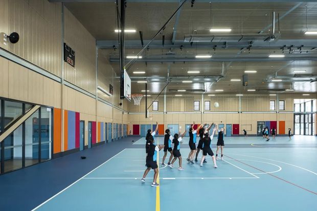 A large indoor multi-purpose sports hall with basketball hoops, marked basketball courts, and players practicing on a pale blue floor. The space has high ceilings, exposed services and lighting, pale timber wall panelling, extensive glazing, and colorful vertical wall panels and doors.