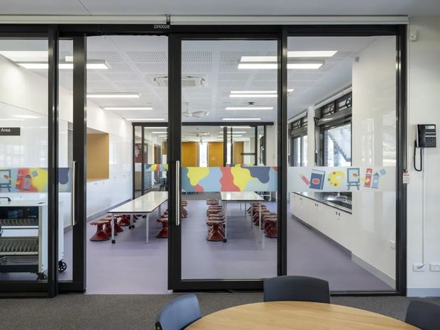 View through black-framed glazed doors into a bright, contemporary educational or commercial interior. The central space contains long tables with small red stools, colourful privacy graphics on the glazing, white built-in cabinetry along the right wall, and acoustic ceiling panels with recessed lighting. A round meeting table and chairs are partially visible in the foreground.