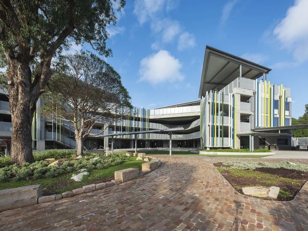 Exterior view of a contemporary multi-storey institutional or commercial building set within landscaped grounds. The building features layered horizontal volumes, extensive glazing, open circulation or balcony zones, prominent vertical fins in varied light colours, and a large projecting roof canopy. A broad brick-paved driveway or forecourt leads toward the entrance, with mature trees and low planting in the foreground.
