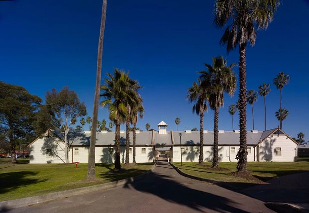 Single-storey light-coloured institutional or commercial building viewed from a paved driveway, with a long pitched roof, repeated rectangular windows, a small central cupola, and a gabled entrance wing. Tall palm trees and manicured lawns frame the symmetrical frontage under a clear blue sky.