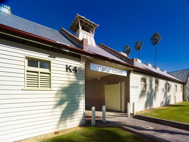 Exterior view of a low-rise heritage-style building clad in pale horizontal weatherboards, with a corrugated metal roof, red roof trim, decorative rooftop cupola, louvred windows, and a recessed entrance marked “Stable Square” and “K4”. The building sits beside a neatly maintained lawn and paved walkway under bright blue skies.