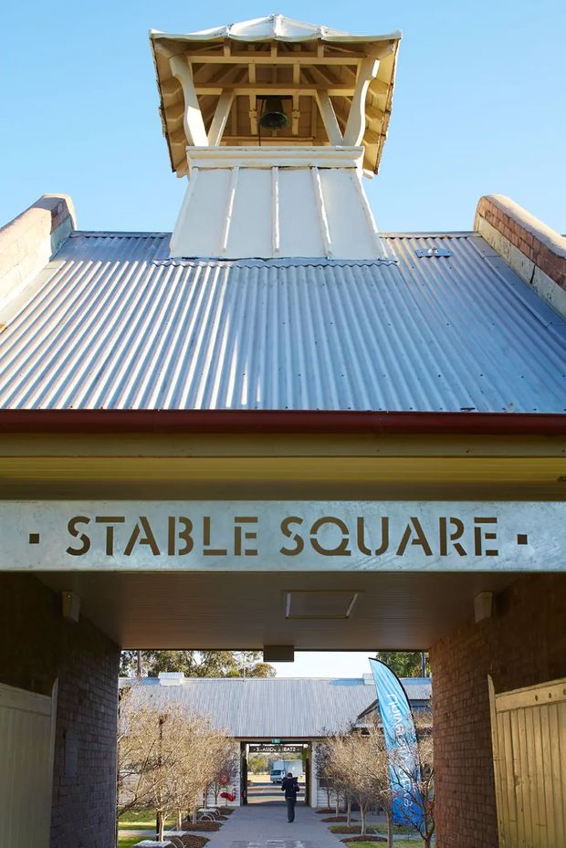 Exterior view of a heritage-style commercial complex entrance labelled “Stable Square.” The image looks upward beneath a corrugated metal roof toward a prominent timber-framed cupola or tower, with brick side walls and a covered pedestrian passage leading into a landscaped courtyard. Bright clear daylight emphasizes the aged white, beige, grey, and brick tones.